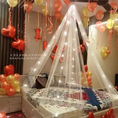 This cozy and romantic bedroom setup features a canopy over the bed woven with fairy lights. The room is filled with red and gold balloons, and 'I LOVE YOU' foil letters are placed by the bed to complete the surprise.