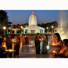 Devotees hold diyas during an evening ceremony outside an illuminated temple. The warm light from the lamps and the joyful expressions capture the beautiful atmosphere of a community united in faith.