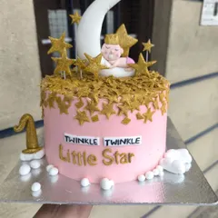 A "Twinkle Twinkle Little Star" themed cake for a baby's celebration. This pink cake is adorned with a sleeping baby on a crescent moon, surrounded by a cascade of golden stars.
