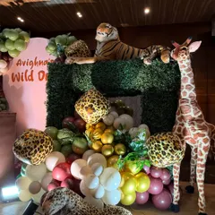 Another angle of the 'Wild One' jungle theme party, showing the arrangement of large stuffed animals like a tiger, giraffe, and leopard around a green hedge prop and balloon display.