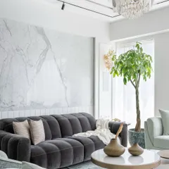 A close-up of a luxury living room where a plush tufted charcoal sofa adds depth against a white marble wall. A statement chandelier and track lighting provide layered illumination, while a large indoor plant brings freshness and vibrancy.