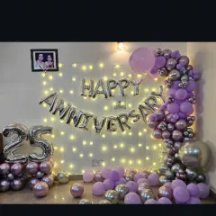 A beautiful 25th-anniversary decoration at home with a fairy light wall, 'Happy Anniversary' foil balloons, and a purple and silver balloon garland.