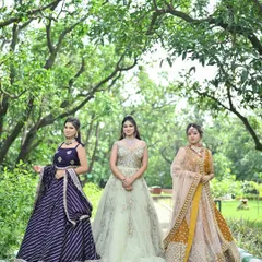 A beautiful outdoor shot of three friends dressed for a wedding. This photo showcases a range of styles available for rent, from a striped purple lehenga to an elegant gown and a traditional mustard lehenga.