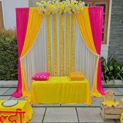 A clean and elegant Haldi stage setup with bright pink and yellow drapes. The backdrop is accented with vertical strings of white flowers, creating a simple yet beautiful space for the ceremony at home.