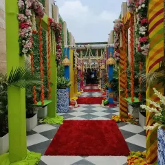 The grand entrance to a festive Teej celebration. The walkway is lined with colorful pillars, traditional jhoolas (swings) decorated with marigolds, and a striking red carpet over a black-and-white checkered floor.