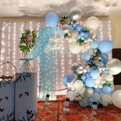 A beautiful birthday party setup with a blue, white, and silver color scheme. The decor includes a large balloon arch, a "Happy Birthday" neon sign, and decorative pedestals, creating an elegant photo corner.