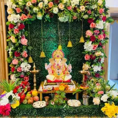 This home Ganpati setup combines a lush green backdrop with a frame of soft-colored roses and traditional bells, creating a fresh and vibrant look.