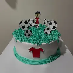 A football-themed cake with green piping to look like grass. This design features a player, footballs, and a jersey made from paper toppers.