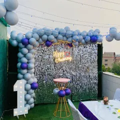 A stylish first birthday party on a rooftop terrace. The decor includes a balloon arch in grey and purple against a silver shimmer wall, a light-up marquee number, and string lights overhead for a festive evening glow.