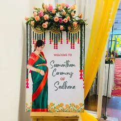 A traditional welcome board for a Seemantham ceremony. The design features an illustration of a mother-to-be in a saree, with marigold and floral garland motifs.