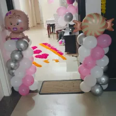 A sweet entrance setup for a baby girl's arrival. Balloon pillars with baby and candy foil balloons stand on either side of a flower petal rangoli, creating a cheerful welcome.