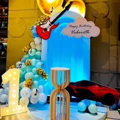 A unique theme combining music and motors for Vedarath's first birthday. The stage features a red sports car, a guitar cutout, musical notes, and cool blue lighting for a stylish celebration.