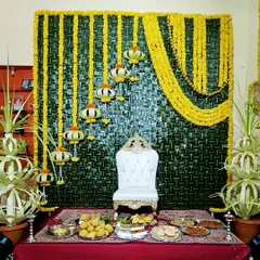 This traditional baby shower setup features a backdrop woven from fresh coconut leaves. The decor is completed with yellow floral arrangements, a special white throne for the mom-to-be, and a table of offerings.