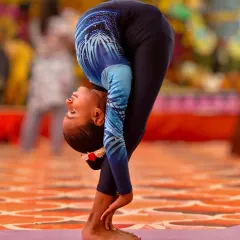 A young student demonstrates a deep standing forward bend, or Uttanasana variation, wrapping her arms for an intense hamstring and spine stretch. This level of flexibility comes from daily, disciplined effort.
