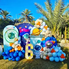 An outdoor space-themed party with a beautiful natural backdrop of palm trees. The setup includes multiple arched panels, astronaut cutouts, and a light-up number '6'.