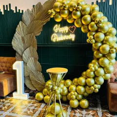 I designed this elegant and modern first-anniversary decoration in a restaurant. The setup features a full ring of metallic gold balloons, dried pampas grass accents, a 'Happy Anniversary' neon sign, and a lit marquee number '1'.