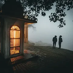 Two figures stand on the edge of a ghat in the deep fog, their silhouettes framed by a small, warmly lit shrine. The image evokes a sense of peace, mystery, and quiet companionship.