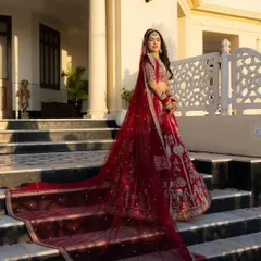 A classic bridal portrait showcasing the bride, Sushmita, in her magnificent red lehenga. The long, flowing veil and regal pose against the palace architecture create a timeless and powerful image of bridal elegance.