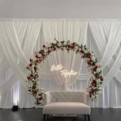 This modern and minimalist engagement setup features a circular arch decorated with red and white roses against a clean white curtain backdrop. The "Better Together" neon sign adds a personal and trendy touch.