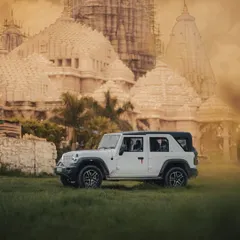 A side profile of the white Mahindra Thar on a lush green field, with the temple architecture in the background. This composition highlights the car's strong presence and versatility, equally at home in both rugged and serene environments.