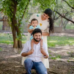 One frame, endless love. A happy baby enjoys a shoulder ride from dad while mom looks on, all surrounded by the beautiful greenery of a park.