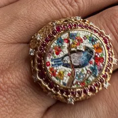 This cocktail ring features a bluebird nestled among blossoms, created with the micromosaic technique. The scene is framed by a halo of vibrant Mozambique rubies and diamonds, set in an intricate 18k gold band.