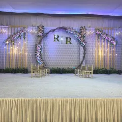 For this engagement, we designed a modern stage with a large central ring featuring the couple's initials. The backdrop includes gold-toned geometric panels and soft floral arrangements, all illuminated by warm bulb lighting.