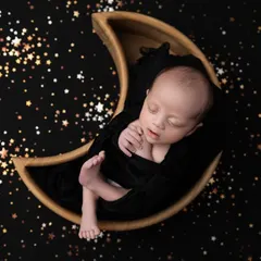 A beautiful contrast of a baby in a dark wrap, nestled in a moon prop against a starry night backdrop. This creates a dramatic and magical celestial portrait.