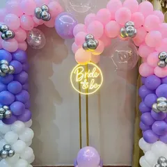 Here is another color option for a bride-to-be balloon arch. This setup uses a mix of pink, purple, and white balloons with silver accents, centered around a "Bride to be" neon sign on a stand for a softer, colorful look.
