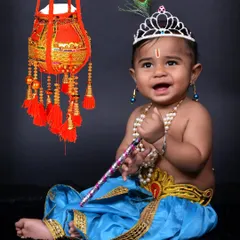 A joyful baby Krishna portrait against a simple dark background. The focus here is on the cute expression and the detailed costume, including the crown and pearl jewelry.