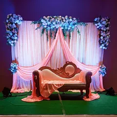 An elegant engagement stage setup with soft peach draping against a darker wall, highlighted by beautiful white and blue floral arrangements and a classic carved sofa for the couple.
