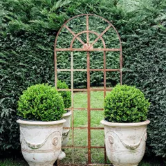 In this garden, we used a rustic arched garden mirror to create an illusion of depth and light. It is framed by two large planters containing perfectly spherical boxwood topiaries, adding a touch of formal symmetry to the lush green backdrop.