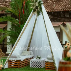 A cozy boho lounge area for a Mehendi. The setup includes a simple teepee tent, a wicker peacock chair, and seating made from wooden crates, creating a relaxed and stylish vibe.