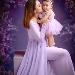 A stunning mother-daughter portrait with a rich, purple floral background. The coordinated lavender outfits and the mother's loving kiss create a vibrant yet intimate fine-art image.