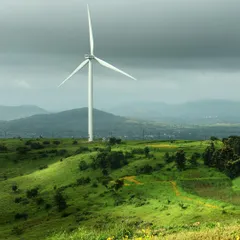 A solitary windmill stands tall over the vibrant green hills, its clean white form contrasting with the moody monsoon sky. It represents the blend of nature and modern technology in the landscape.