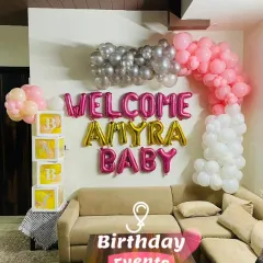 We can transform your living room into a festive space for your baby's arrival. This setup for 'Amyra' includes a custom balloon arch in pink, white, and silver over the sofa, along with our popular transparent 'B-A-B-Y' boxes.