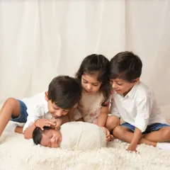 Three older siblings gather around their new baby sibling, looking down with curiosity and love. One gently touches the baby's head, marking the start of their bond.
