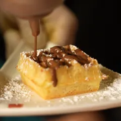 Pure indulgence in the making. A freshly made waffle square gets a generous drizzle of rich chocolate sauce, promising a warm, sweet, and satisfying dessert experience.