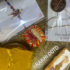 The perfect Rakhi OOTD (Outfit Of The Day) setup. This flat-lay shows our unique rakhis, including a Superman design and a "No Pain No Gain" lapel pin, ready for the celebration with traditional sweets.