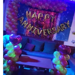A cozy anniversary decoration in a dimly lit room. The setup includes a balloon arch, 'Happy Anniversary' foil letters, and heart-shaped foil balloons.