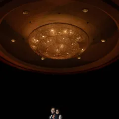 A dramatic portrait using negative space, with the couple positioned under a massive, ornate chandelier. The composition emphasizes the grandeur of the location and the intimacy of the couple.
