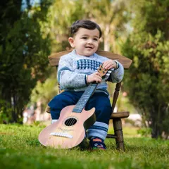A toddler boy sits on a small wooden chair in a park, holding a guitar and smiling. A natural and joyful outdoor portrait.