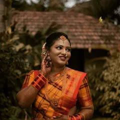 This portrait, taken at a rustic outdoor location, captures a candid moment of reflection. The warm, natural light complements the beautiful yellow and red checked saree, highlighting the intricate gold jewelry and the bride's serene expression.