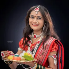 A bright and cheerful portrait of a woman in a red and black saree, holding a decorative puja thali for a Sankranti themed shoot. Her look is completed with traditional sugar jewelry.