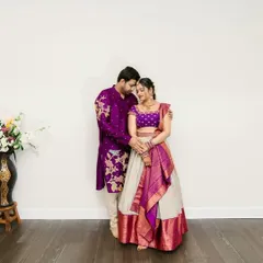 A full view of the couple, showcasing the bride's elegant ivory and purple half-saree lehenga and the groom's matching embroidered kurta.