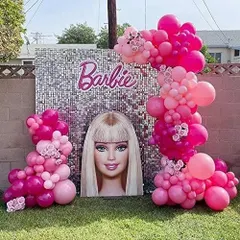 A glamorous outdoor Barbie-themed party. The setup includes a silver shimmer wall, a large Barbie head cutout, and beautiful pink organic balloon garlands.