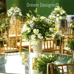 A beautiful mirrored tabletop reflecting a classic centerpiece. The arrangement of white lilies and roses is set in a pearl-covered vase, with pearl strings draping from the chairs to complete the elegant, pearlescent theme.