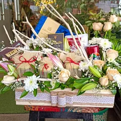 A beautiful birthday hamper that combines rustic charm with floral elegance. It features a large arrangement of white and peach roses, complemented by gourmet chocolates and snacks.