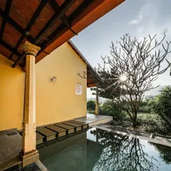 The sun shines through a bare frangipani tree, casting beautiful reflections on the private pool of a villa at Dvara Resorts. The traditional stone pillar and yellow wall create a timeless and serene atmosphere.