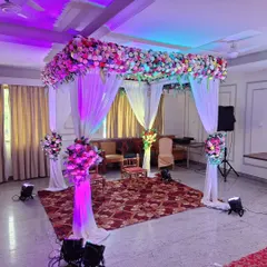 This is a four-pillar mandap setup for an indoor engagement or wedding ceremony. The canopy is heavily decorated with multi-colored flowers, and the pillars are draped in white fabric with matching floral bouquets.
